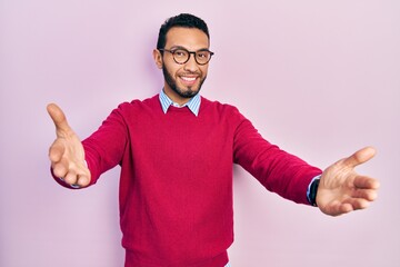 Hispanic man with beard wearing business shirt and glasses looking at the camera smiling with open arms for hug. cheerful expression embracing happiness.