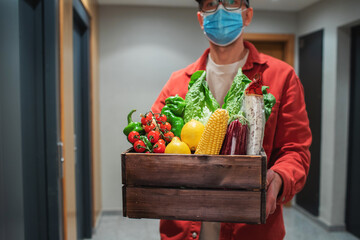 Delivery man in protective mask holding paper bag with food in the entrance. The courier gives the box with fresh vegetables and fruits to the customer