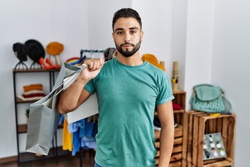 Young handsome man with beard holding shopping bags at retail shop relaxed with serious expression on face. simple and natural looking at the camera.