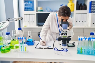 Young hispanic woman wearing scientist uniform using microscope and writing on clipboard at laboratory