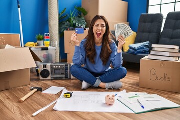 Young brunette woman sitting on the floor at new home holding savings winking looking at the camera...