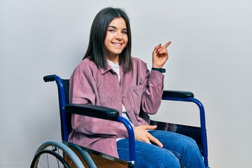 Young brunette woman sitting on wheelchair with a big smile on face, pointing with hand finger to the side looking at the camera.