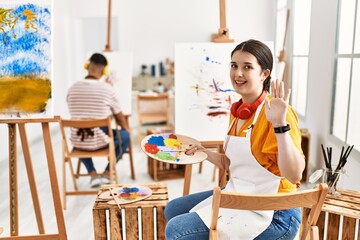 Young artist woman painting on canvas at art studio waiving saying hello happy and smiling,...