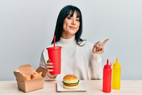 Young brunette woman with bangs eating a tasty classic burger with ketchup and mustard smiling happy pointing with hand and finger to the side