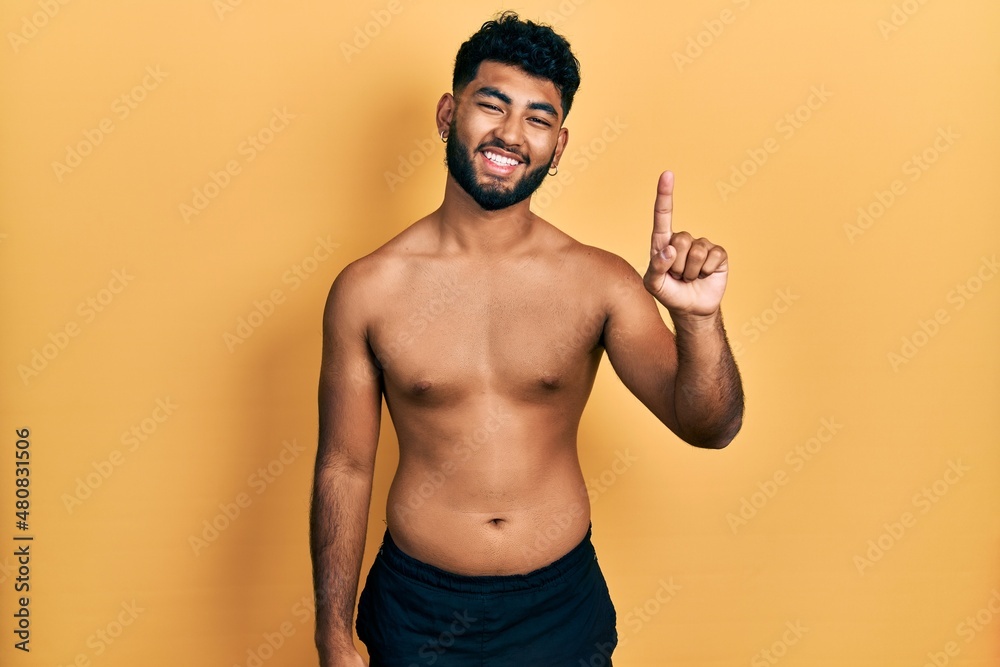 Poster arab man with beard wearing swimwear shirtless showing and pointing up with finger number one while 