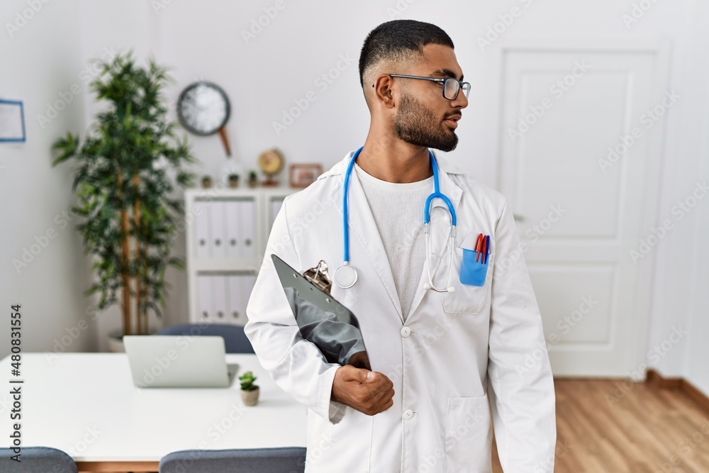 Sticker Young indian man wearing doctor uniform and stethoscope looking to side, relax profile pose with natural face and confident smile.