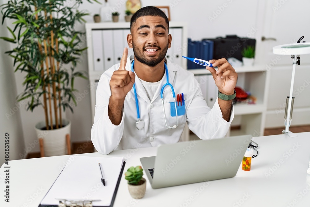 Wall mural Young indian doctor holding thermometer smiling with an idea or question pointing finger with happy face, number one