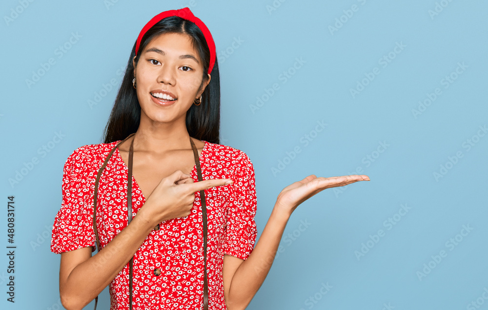 Poster Beautiful young asian woman wearing dress and vintage camera amazed and smiling to the camera while presenting with hand and pointing with finger.