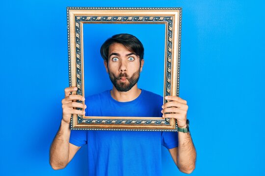 Young hispanic man holding empty frame making fish face with mouth and squinting eyes, crazy and comical.