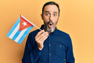 Middle age hispanic man holding cuba flag scared and amazed with open mouth for surprise, disbelief...