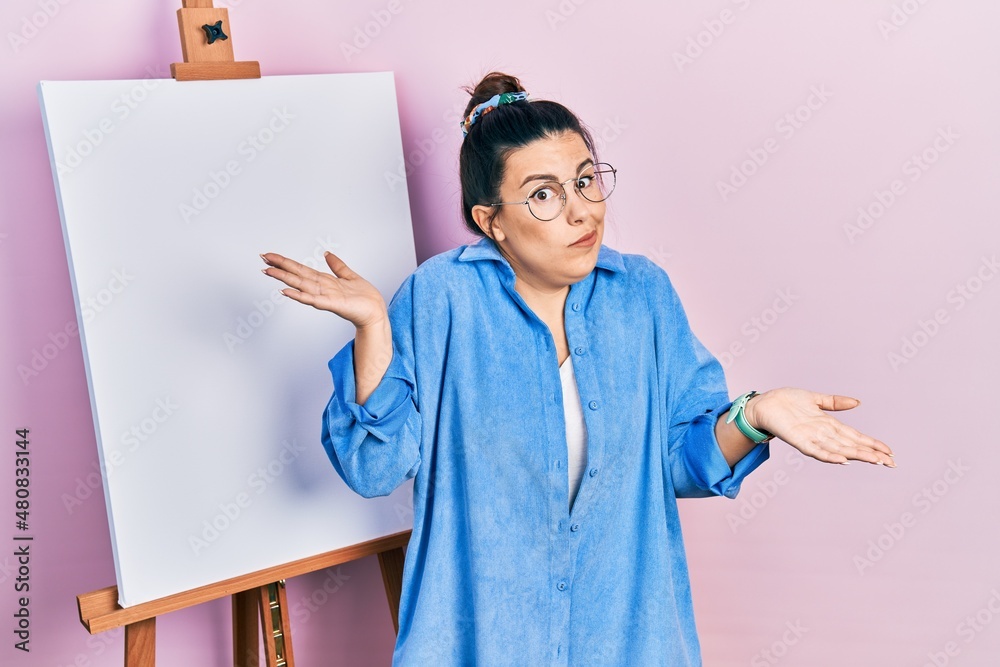 Sticker Young hispanic woman standing by painter easel stand clueless and confused expression with arms and hands raised. doubt concept.