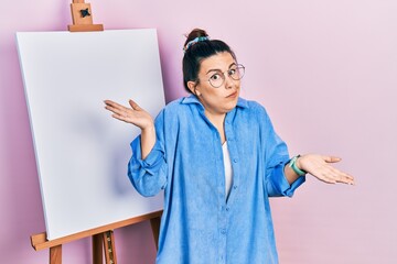 Young hispanic woman standing by painter easel stand clueless and confused expression with arms and...