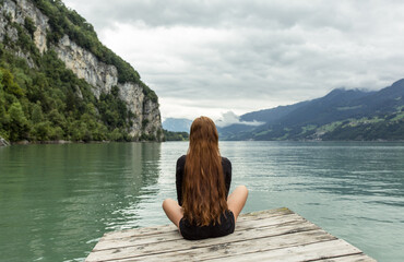 Relaxed girl sitting alone meditating on a dock facing the beautiful lake view. 
