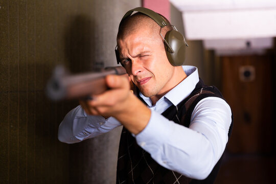 Focused man in ear protectors aiming single barrelled shotgun at target in shooting range.