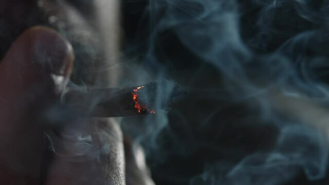 Fingers holding a marijuana joint, smoking it in crime with pleasure while cigarette burning and turning into ash, close-up on the black background.