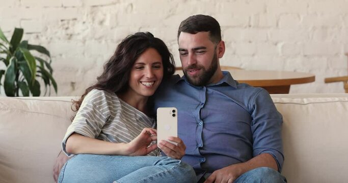 Cheerful couple sit on couch, use cellphone read social media news look overjoyed, receive great commercial offer, buying online feel happy, enjoy easy remote comfort electronic service usage concept