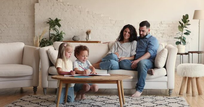 5s kids drawing with pencils sit at table, their parents relax on sofa, family gather in cozy modern living room spend weekend leisure enjoy rest and hobby. Happy homeowners family, upbringing concept