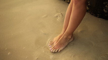 View top, from above. The woman feet relaxed are stand on the sandy beach and washed by the water and foam of the ocean