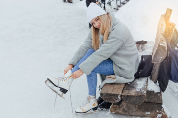 Lovely young woman sitting on the bench and puting on figure skates on the ice rink.