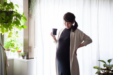 モーニングコーヒーを飲みながら窓辺に立つ、妊婦の女性