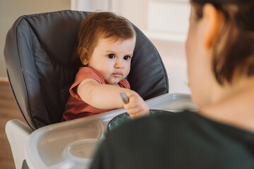 Messy smiling baby girl eating with a spoon in high chair. Natural organic nutrition. Organic food. Baby care.