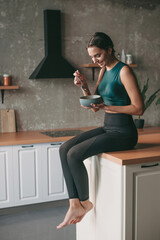 Caucasian woman in sportswear resting at the kitchen after exercise and eating a bowl of porridge....