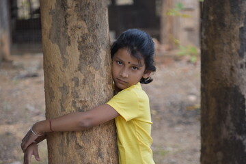A girl kid holding tree trunk with her arm normal expressions wearing yellow tshirt selectively focused blur background 