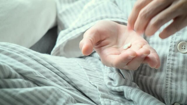 A view focused on the hand of a sick and exhausted woman dressed in white, pouring a pill of medicine from a pill bottle, putting it on her hand and taking it to relieve her sickness.