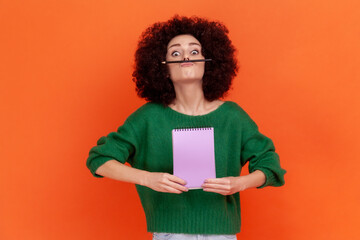 Funny young adult woman with Afro hairstyle wearing green sweater having fun holding pencil with lip and nose, female student resting during hometask. Indoor studio shot isolated on orange background.