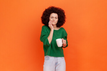 Sad sick woman with Afro hairstyle wearing green casual style sweater has teeth pain after drinking hot or cold beverage, dental injury. Indoor studio shot isolated on orange background.