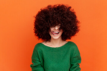 Happy positive woman with Afro hairstyle wearing green casual style sweater showing tongue out, demonstrating her hair, need cutting bangs. Indoor studio shot isolated on orange background.