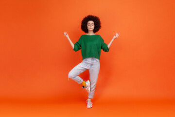 Fototapeta na wymiar Full length portrait of woman with Afro hairstyle wearing green sweater with mudra gesture hands up, closed eyes, meditating standing in yoga position. Indoor studio shot isolated on orange background