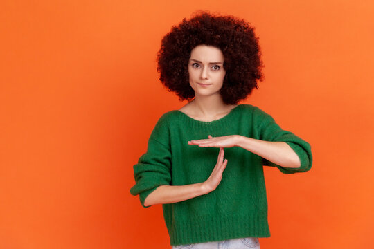 Young adult woman with Afro hairstyle wearing green casual style sweater showing time out hand gesture, looking at camera, worried about deadline. Indoor studio shot isolated on orange background.