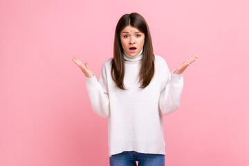 Brunette woman raising hands and looking with confused expression, quarreling, annoyed by conflict, wearing white casual style sweater. Indoor studio shot isolated on pink background.