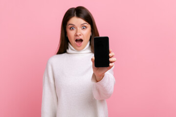 Astonished brunette female showing cell phone with empty display with copy space for advertisement, wearing white casual style sweater. Indoor studio shot isolated on pink background.