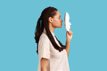 Side view portrait of woman with black dreadlocks holding white mask in front her face, having strict facial expression, wearing white shirt. Indoor studio shot isolated on blue background.