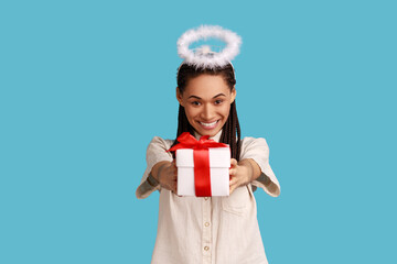 Portrait of cheerful woman with black dreadlocks and halo overhead holding gift box, charity organization, religion, wearing white shirt. Indoor studio shot isolated on blue background.