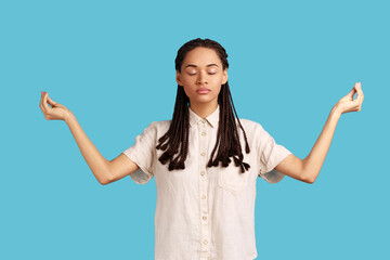Calm relaxed woman with black dreadlocks standing with closed eyes, meditates and enjoys peaceful atmosphere, makes mudra gesture, wearing white shirt. Indoor studio shot isolated on blue background. - obrazy, fototapety, plakaty