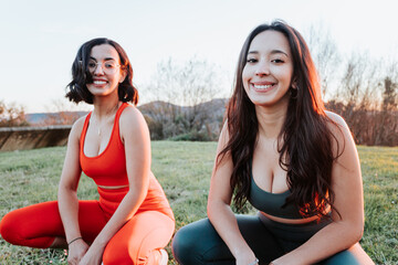Two young african females resting while squatting at the grass during a colorful sunset, portrait one in focus. Training at the park after work, workout at the city, having fun together doing exercise