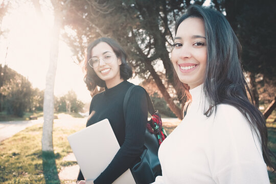 Two young students standing outdoors holding laptop, backpack and study accessories while waiting for the bus to come and take them home. Studying outside home concept. Back to campus