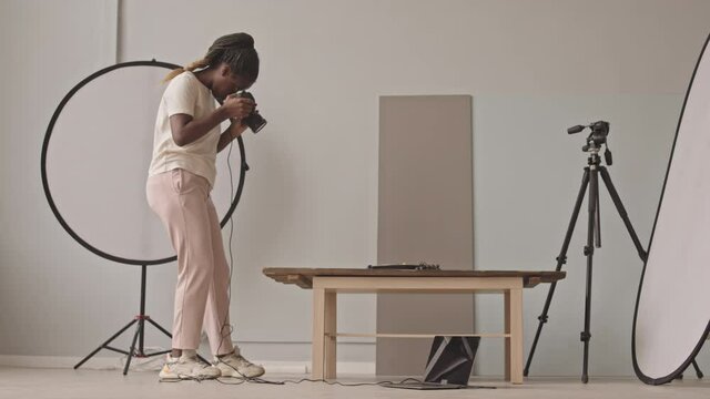 Full-length stab shot of young African-American female photographer taking photos on digital camera in photo studio with professional lighting and equipment