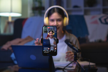 Cheerful beautiful Asian woman wearing headphones and live streaming on a smartphone at home.
