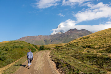 Fototapeta na wymiar A woman walking on a panoramic hiking trail leading to the Koruldi Lakes in the Greater Caucasus Mountain Range in Georgia, Upper Svanet Region. Panoramic view on the Ushba mountain peaks. Wanderlust.