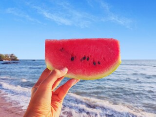Holding a slice of watermelon with blue sky and beach background