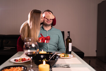 Couple in love celebrating valentine's day at home, girl gives a gift to a man, kisses him with her hand covering her eyes, date of lovers at dinner