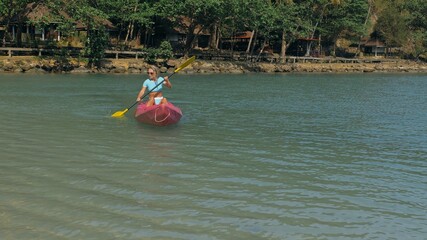 Young blonde woman in blue swimsuit rows pink plastic canoe along azure sea bay past island with palms under blue sky at resort.