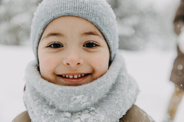 Closeup portrait of a little boy in knitted beanie with a scarf, smiling, looking at camera, on cold winter day, with frost on eyelashes.