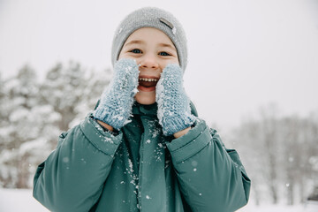 Child on snowy winter day, laughing, covering face with knitted mittens.
