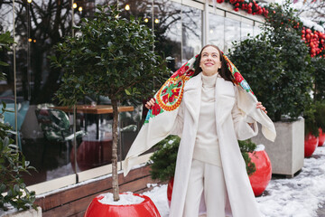 A young girl in winter on the street in a white fur coat and a bright scarf with a crest. Winter style, fashion.