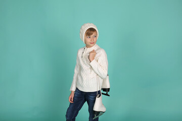 a blue-eyed boy in a white knitted sweater and a winter hat holds figure skating skates in his hands
